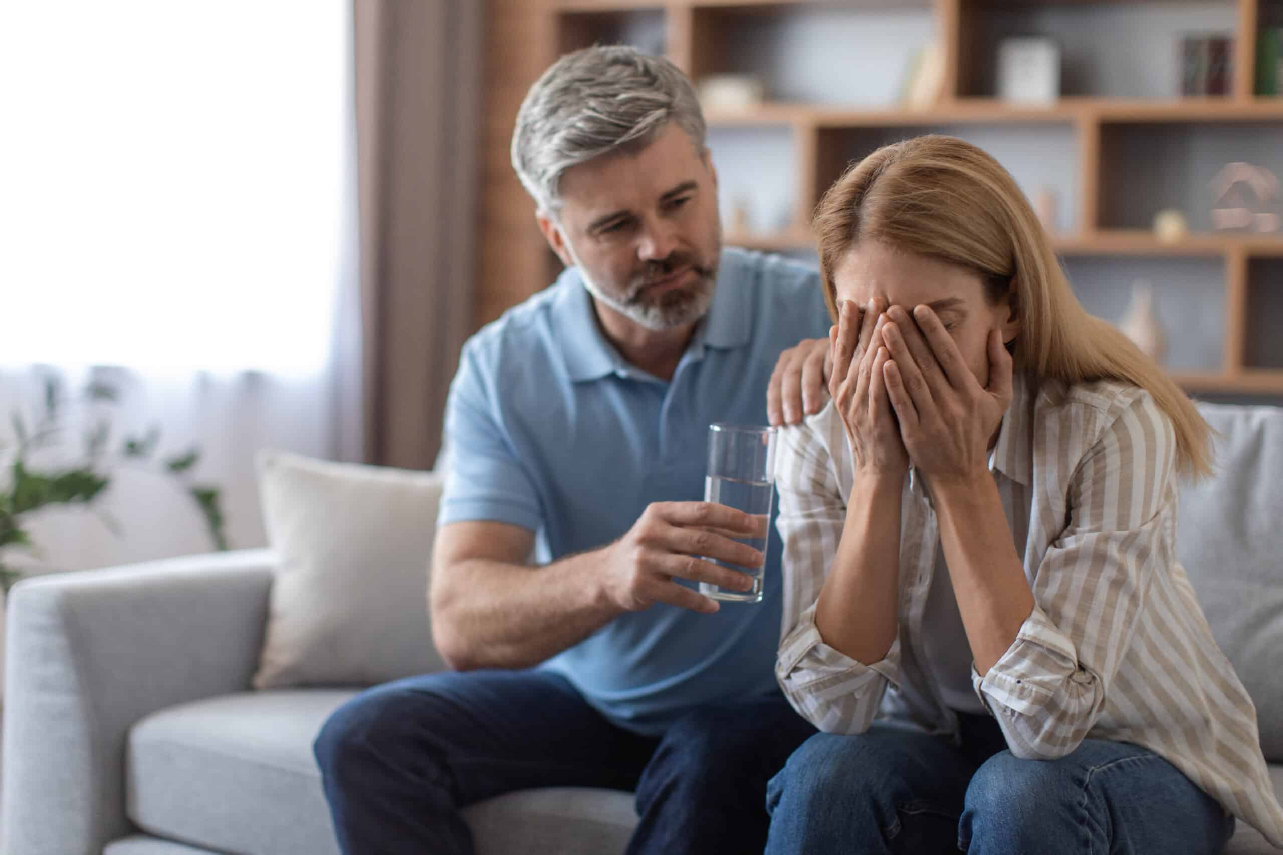 Serious mature caucasian husband calm upset crying wife, gives glass of water, lady suffers from pain and depression in room interior. Bad news, illness, stress, relationship problems and support