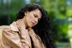 Worried young woman in a stylish beige jacket outdoors, rubbing neck, showing discomfort and pain, with a blurred green background.