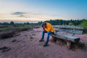 A sad older male clutching his head in his hands on an old wooden bench on a rural field